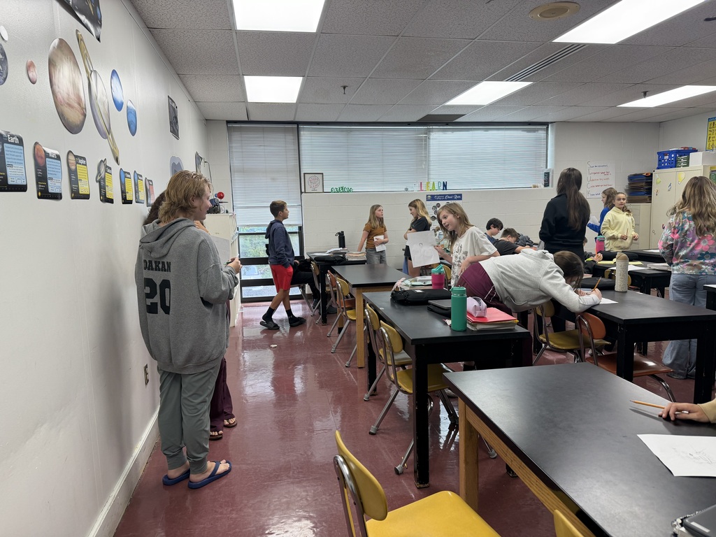 Students standing in a classroom. There are space items on the wall and there are windows in the far end of the room. There are black tables and the lights are on.