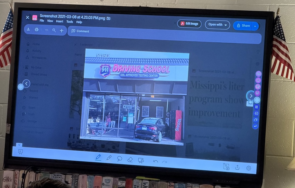 A whiteboard with a photo of a car that has wrecked into a Driving School Center. There are American flags on both top corners of the whiteboard.