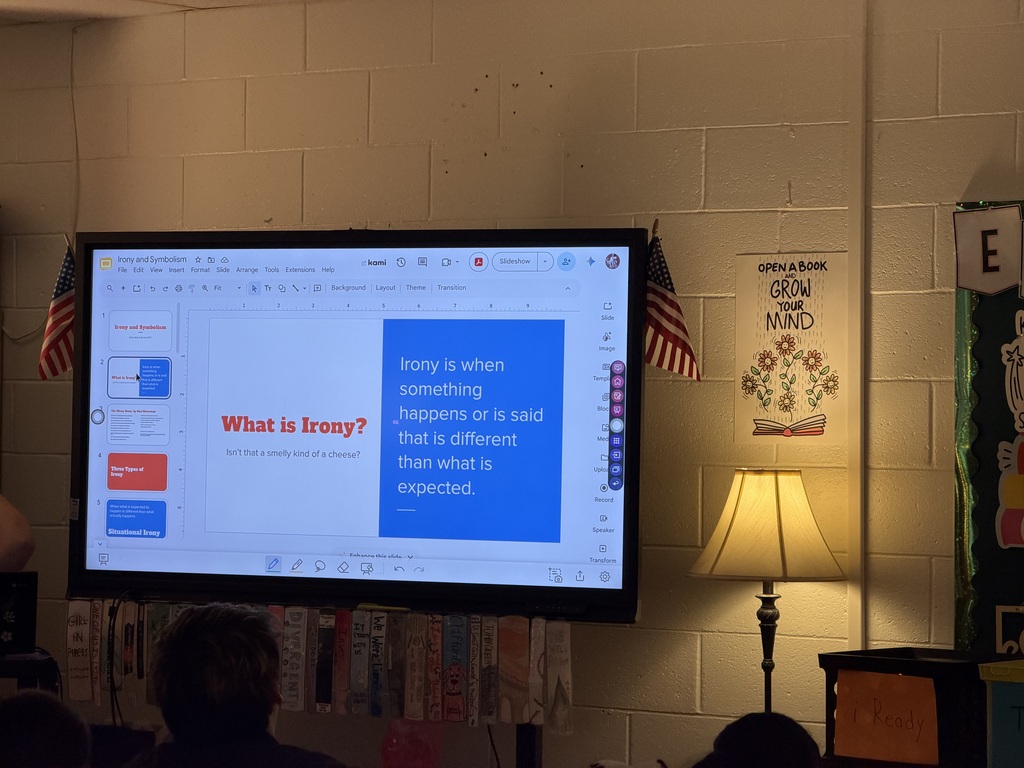A whiteboard in a classroom that says - What is Irony? There is a lamp on beside the whiteboard and there is a sign hanging on the wall with flowers on it that says Open a book grow your mind.