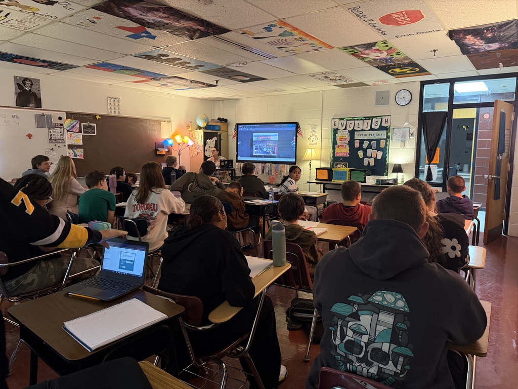 Students sitting at desks in a classroom. A teacher is sitting beside a whiteboard in the front of the class. The classroom door is open and there is a bulletin board in the front of the room that says ENGLISH on it. There are painted ceiling tiles in this classroom.