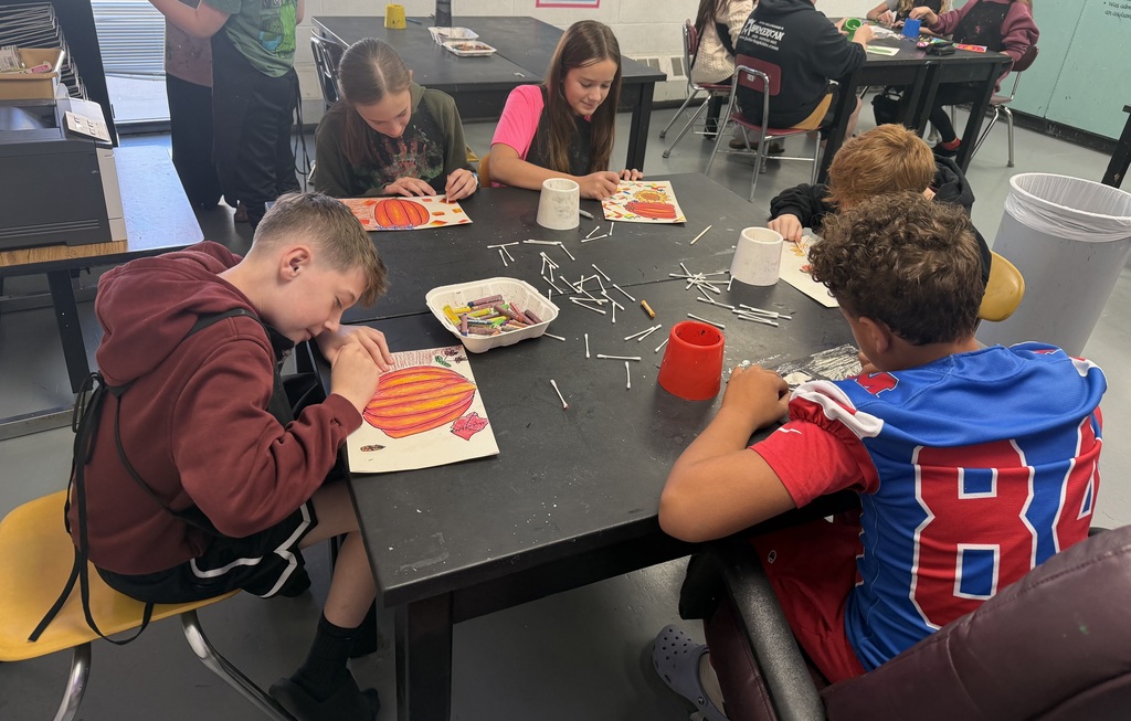 Five students sitting at a black table with oil pastels, paper, and q tips on it. The are coloring fall pictures. One student has a #83 football jersey on.
