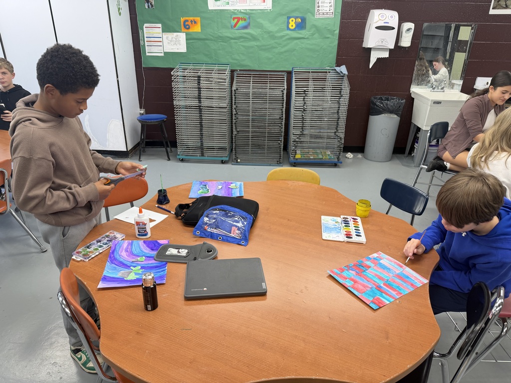Two students are at a table. One is painting and the other one has scissors in his hand. The table has paper, a Chromebook, pencil pouch, Chromebook bag, glue, and a Yonder punch sitting on it. In the background you can see a drying rack, bulletin board, and other students sitting at another table.