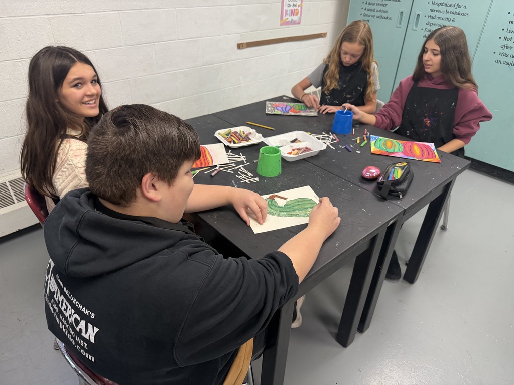 Four students sitting at a black table with paint, q tips, a pencil bag, and papers on it. In front of each of the students is a piece of paper that has fall themed drawn items that have been painted using oil pastels.