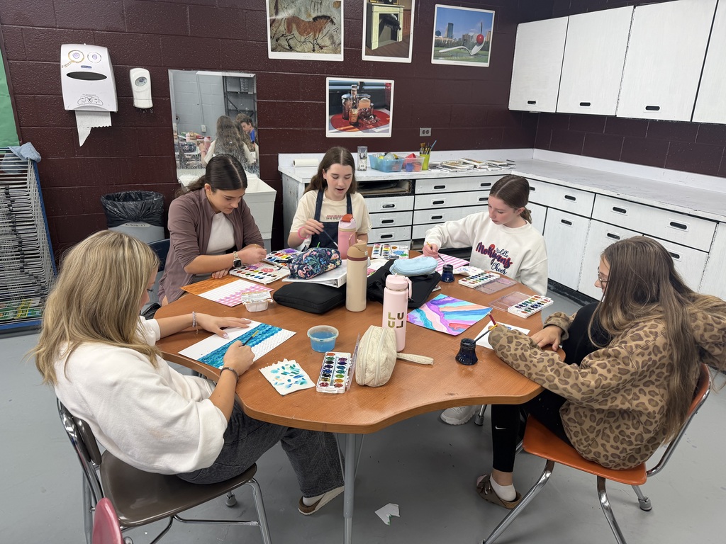 A group of students sitting at a table in an art room. On the table there are paints, papers, and water bottles. All the students are painting on paper. In the background there is a white countertop and cabinets. There is a paper towel holder and 4 art pieces are hanging on the wall.