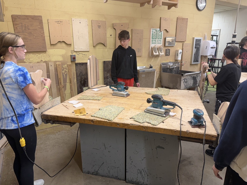 Students are standing at a table working with wood in a workshop setting.