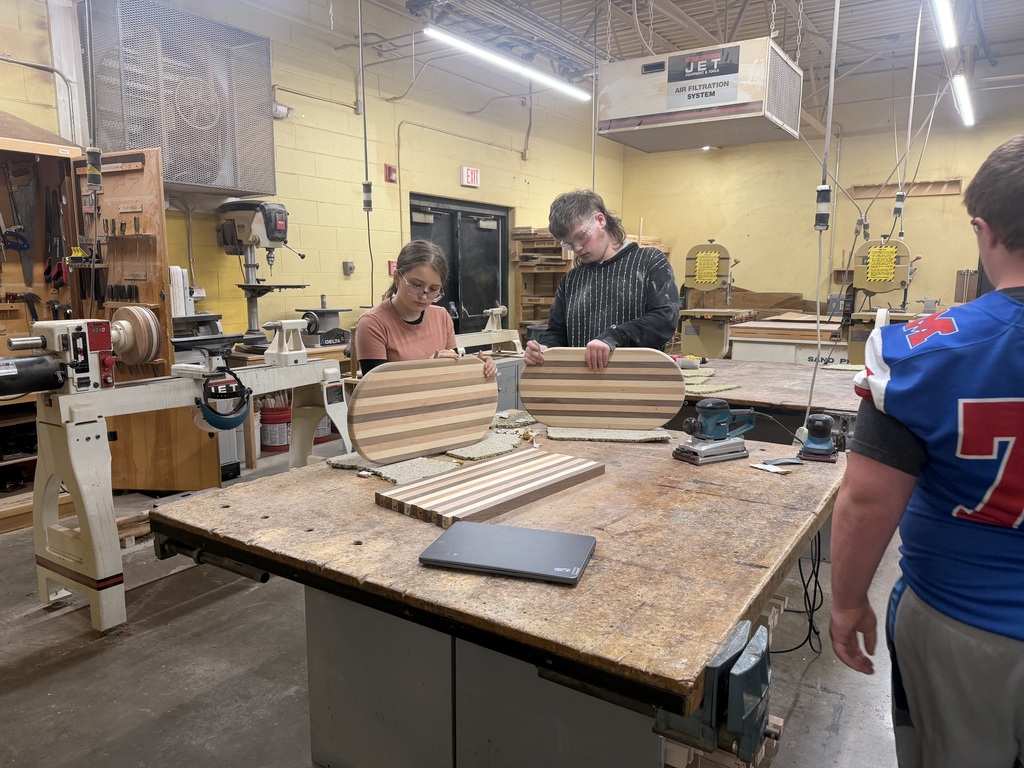 Three students working on creating cutting boards. Two students are sanding their cutting boards. They are all in a workshop.