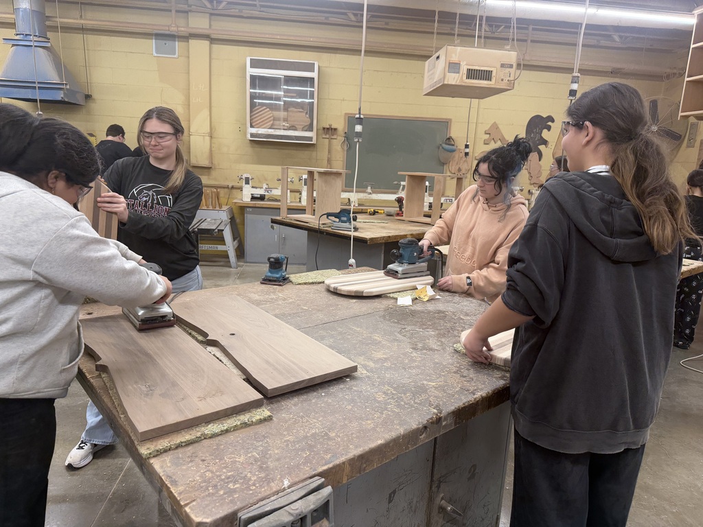 Four students standing around a table in a workshop sanding wood. They are all  wearing safety glasses
