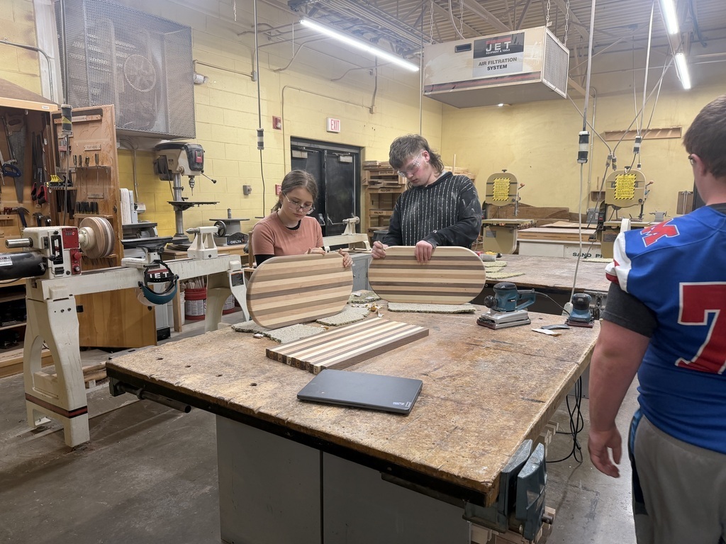 Three students working on creating cutting boards. Two students are sanding their cutting boards. They are all in a workshop.