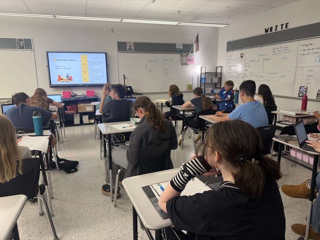 Students with their backs to the camera sitting at desks in what appears to be an ELA classroom. There is a whiteboard in the front of the room with a slide on it. Students have papers on their desks.