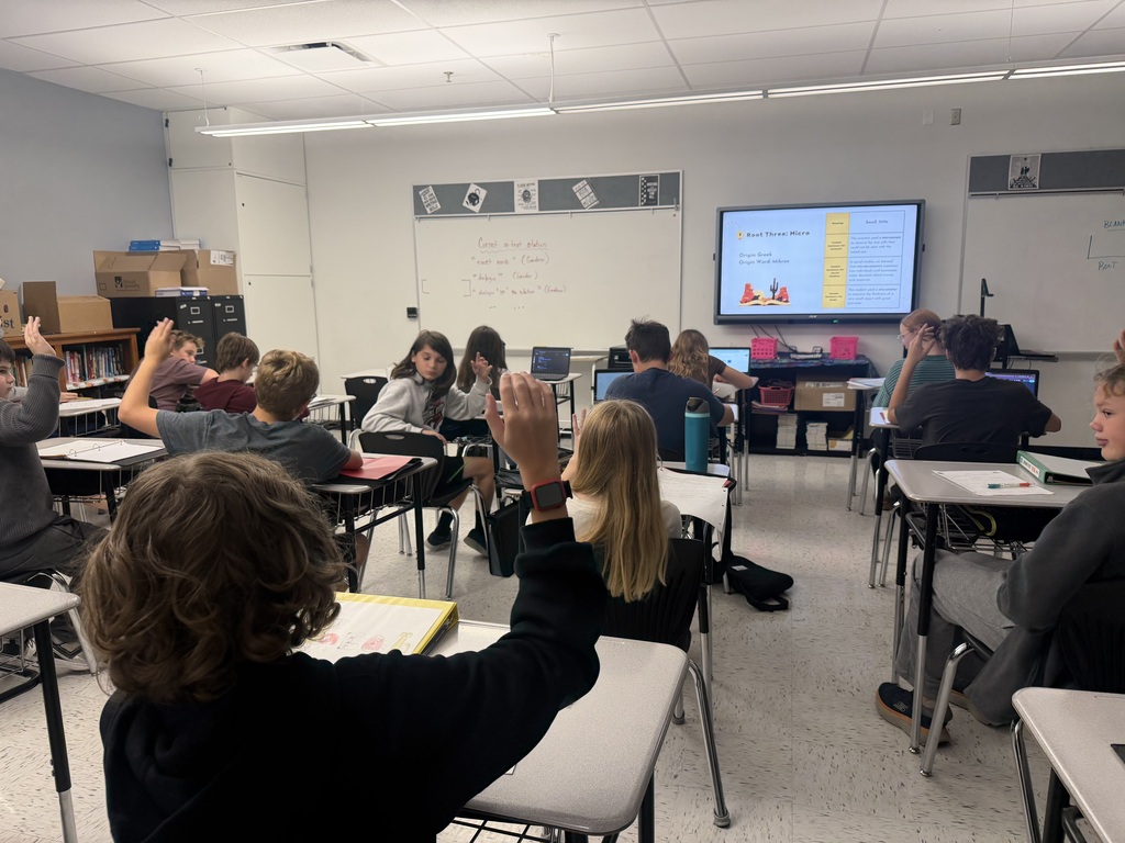 Photo of students sitting at desks, some have their hands up. It appears to be a classroom and there is a whiteboard with a slide on it in the front of the room.