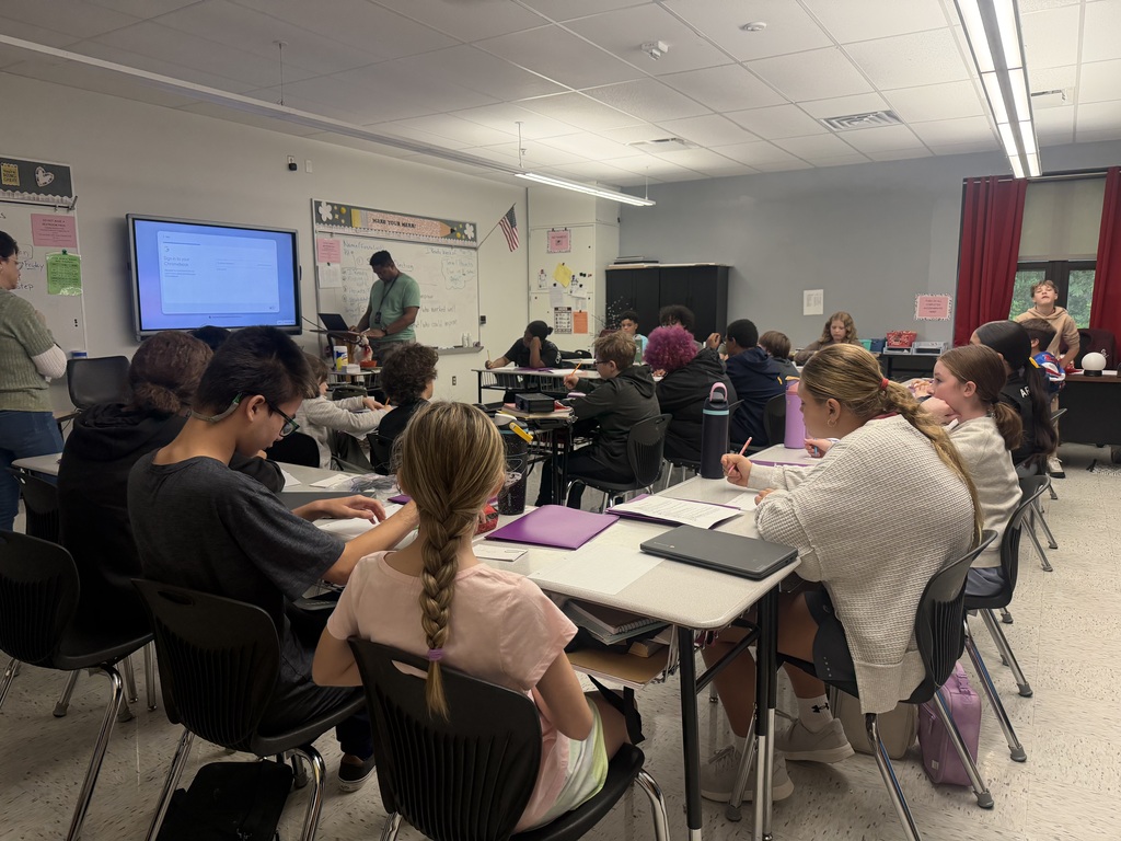Photo of a classroom with students sitting at desks. There is a man in the center of the room looking at a Chromebook. There is a whiteboard at the front of the room