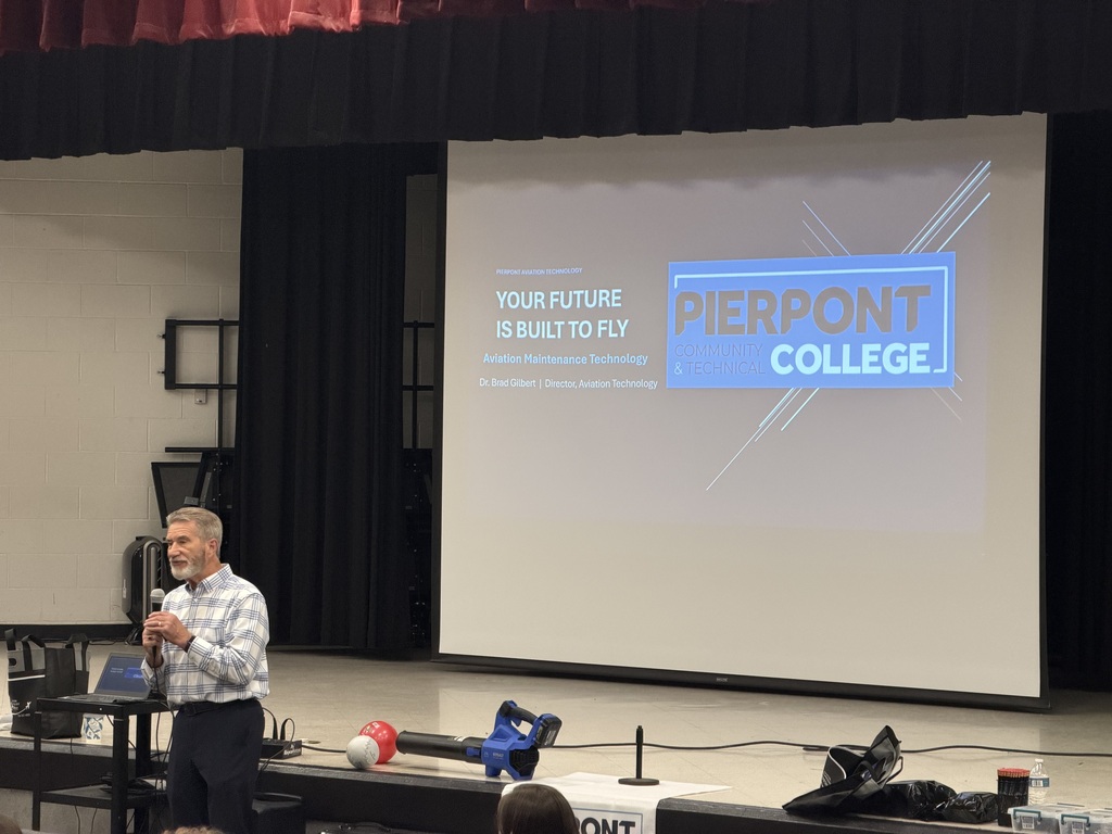 A man with a microphone standing in front of a stage with a screen down. The screen says Your future is build to fly, Pierpont College.