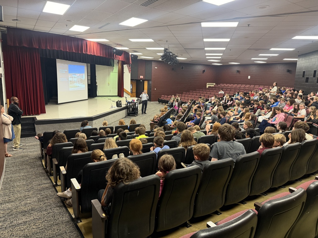 A photo of students sitting in seats in an auditorium setting. There is a man beside a stand in the front of the auditorium with a stage with a screen down. You can see something is projected on the screen. There are a few adults standing along the sides of the walls.