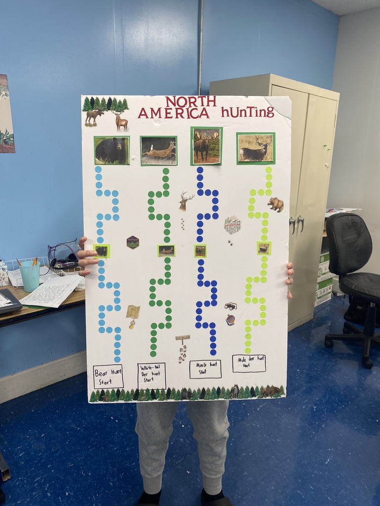 A photo of a person standing in what appears to be a classroom holding a pasteboard that says North America Hunting. There are also a bunch of dots on it that make a path to a box at the bottom of the poster board.