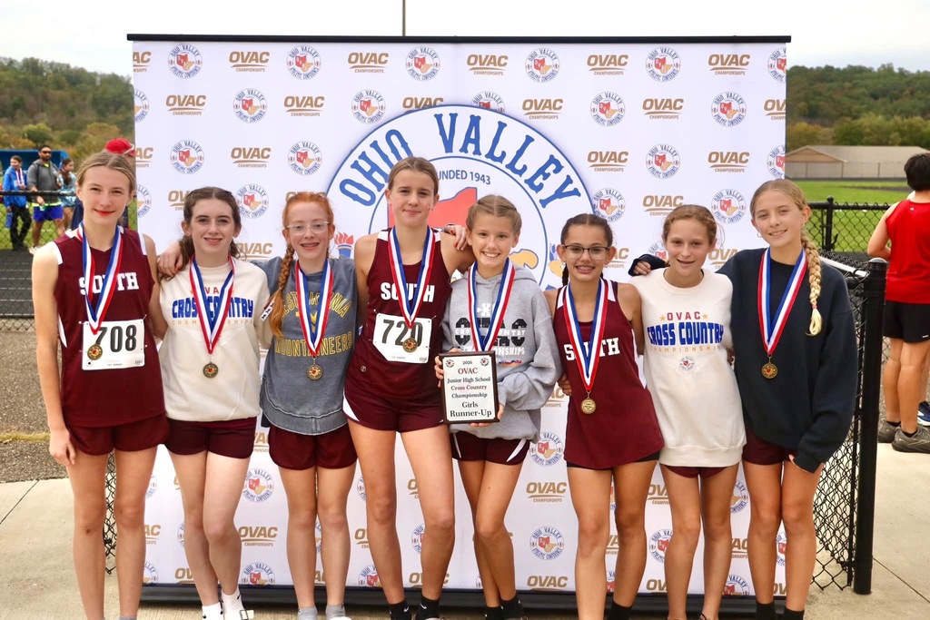 A photo of 8 cross country girl runners standing infant of an Ohio Valley Sign. Each girl is wearing a medal and one is holding a plaque.