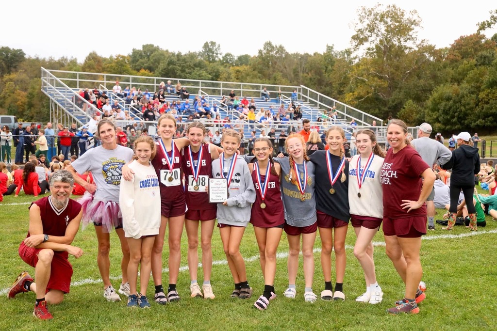 A photo of 8 cross country girl runners. 7 of the runners are wearing medals and one is holding a plaque. There are 3 adults in the photo, one is kneeling on the ground and the other 2 adults are standing. There is a bleacher in the background.