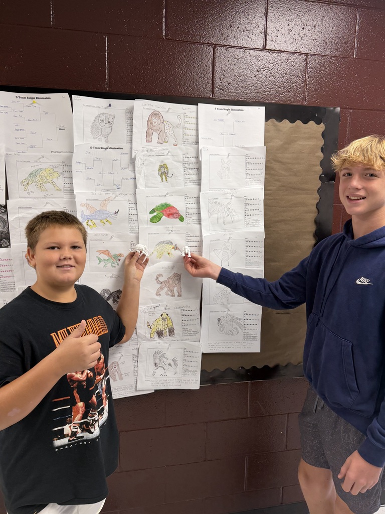 A photo of two students standing in front of a bulletin board with monster drawings on it. The students are each holding up a 3D printer monster.
