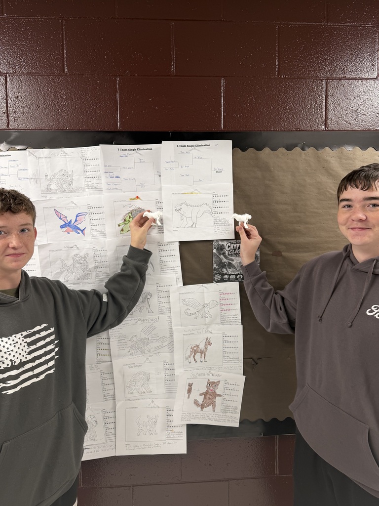 A photo of two students standing in front of a bulletin board with monster drawings on it. The students are each holding up a 3D printer monster.