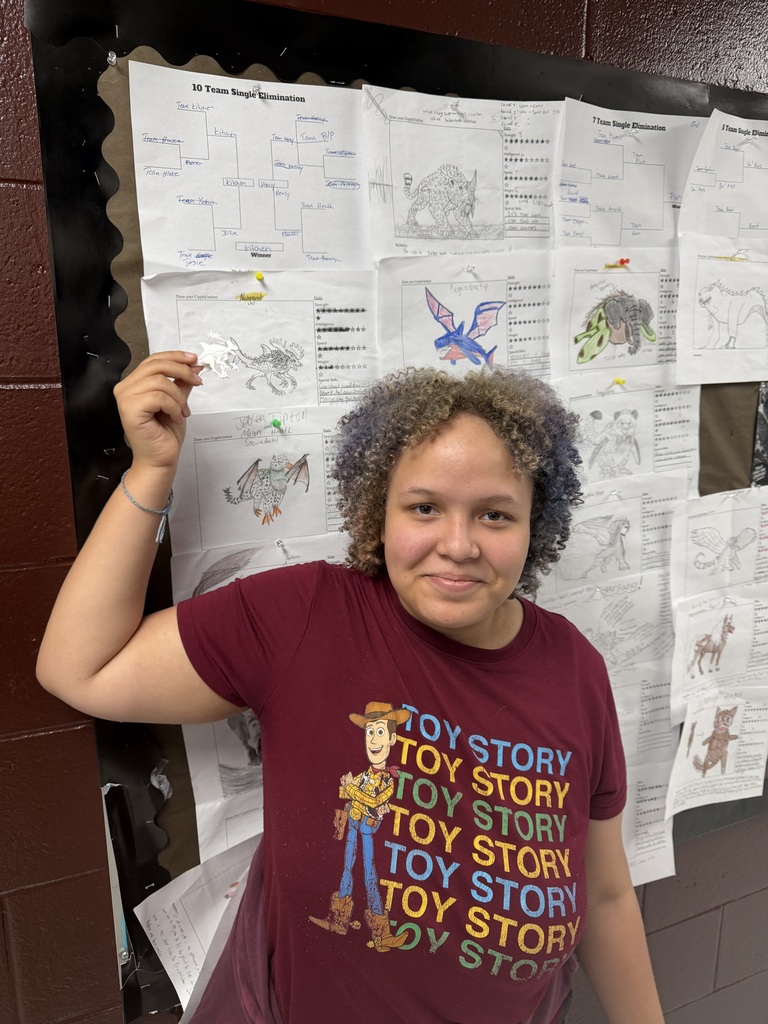 A photo of a student standing in front of a bulletin board with monster drawings on it. The student is holding up a 3D printer monster.