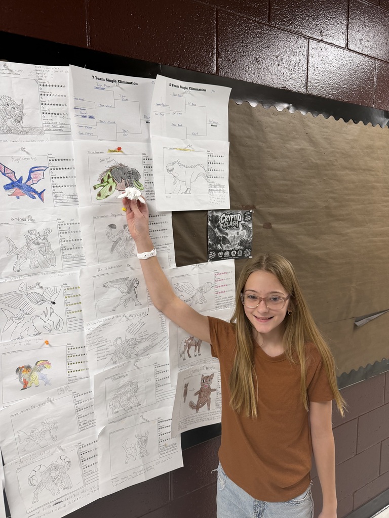 A photo of a student standing in front of a bulletin board with monster drawings on it. The student is holding up a 3D printer monster.