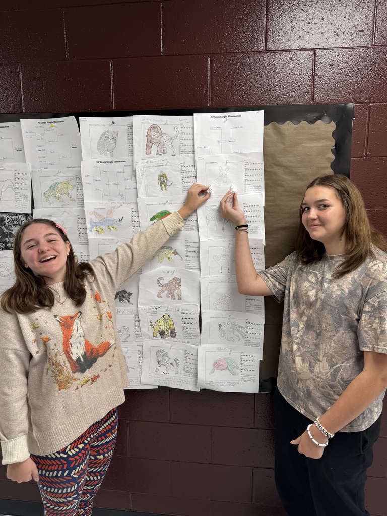 A photo of two students standing in front of a bulletin board with monster drawings on it. The students are each holding up a 3D printer monster.