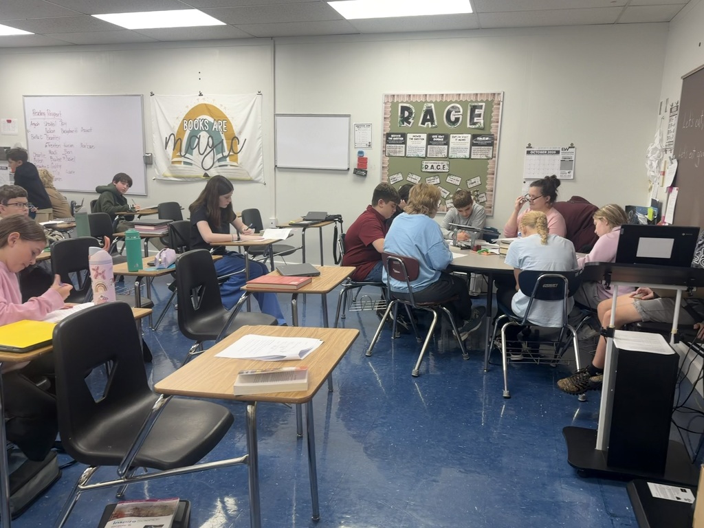 A photo of a classroom where students are working at their desks and a group is working at the table with a teacher