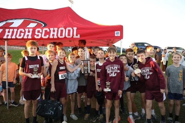 A photo of a group of male cross country runners wearing South CC clothing. They are holding trophies and standing in front of a High School canopy.