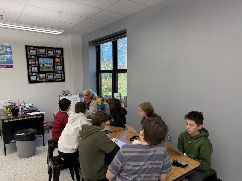 A photo of students sitting at a table with a teacher holding a whiteboard.