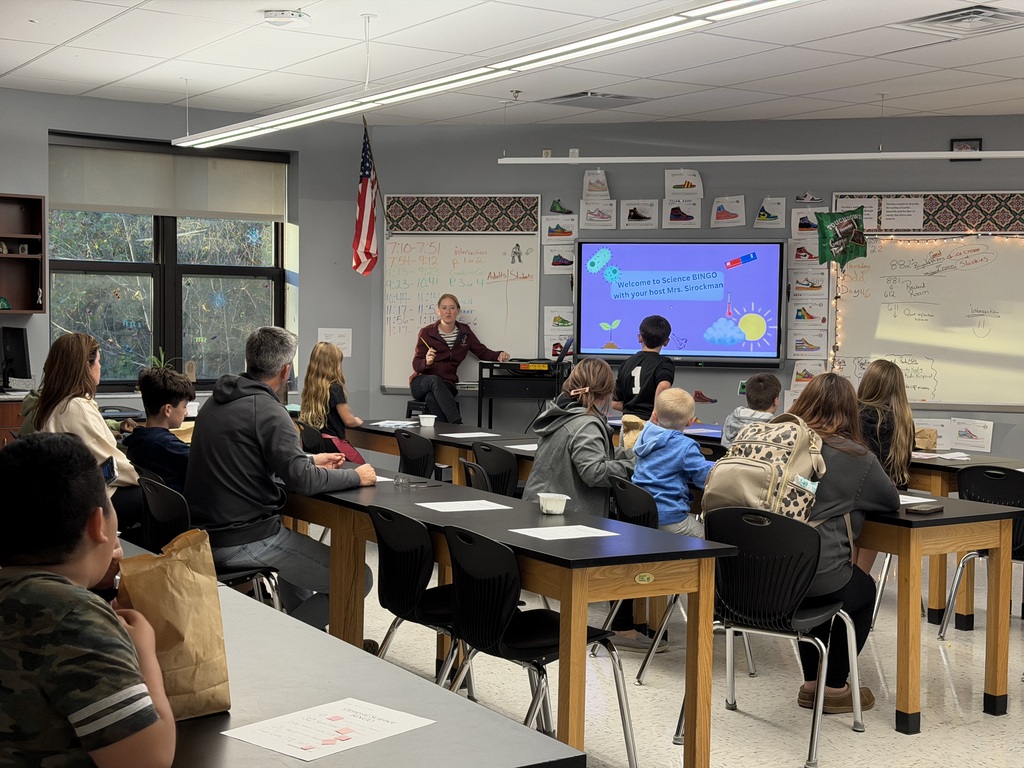 A photo of a classroom filled with parents and students sitting at tables. A woman is sitting on a stool in the front of the room with a whiteboard on. The people in the room are turned towards the woman. It appears like she is telling them something.