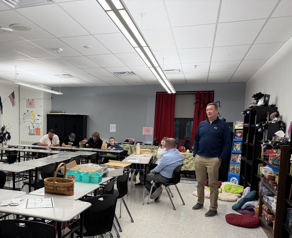 A photo of a classroom where 6 people are sitting and working at desks and one adult is standing.