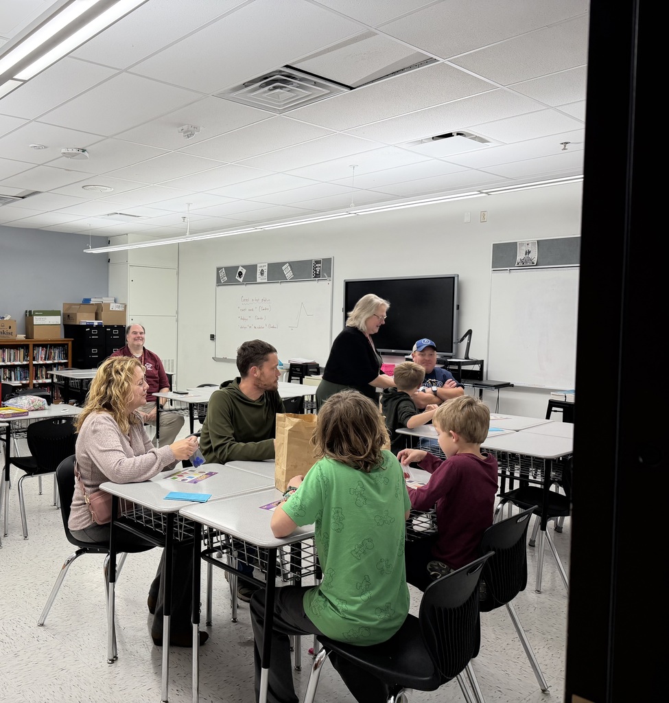 A photo of adults and children sitting at desks in a classroom. It appears if there are BINGO boards on the desks. A woman wearing black is standing by an adult sitting at a desk.