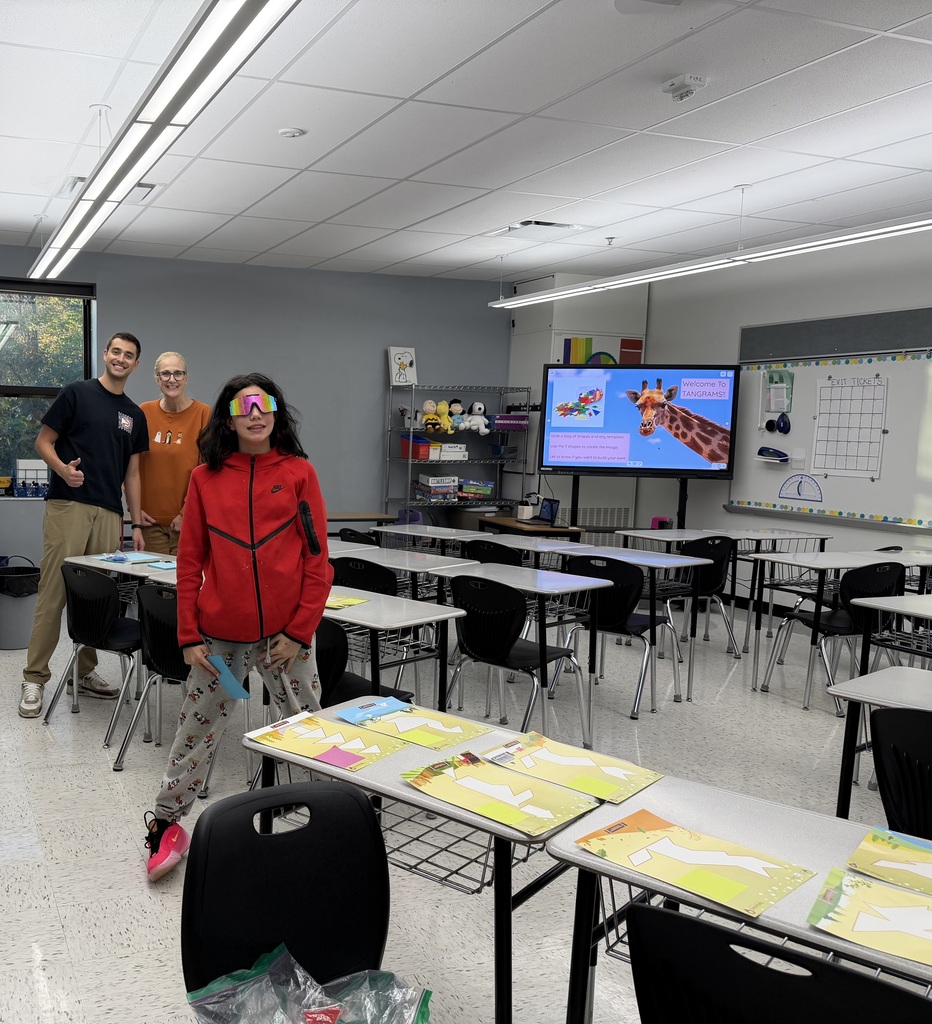 A photo of two adults standing in the back and a teenagers standing in front of them. They are all in a classroom. There are yellow papers on desks in this classroom and a TV is on with a giraffe on the screen.
