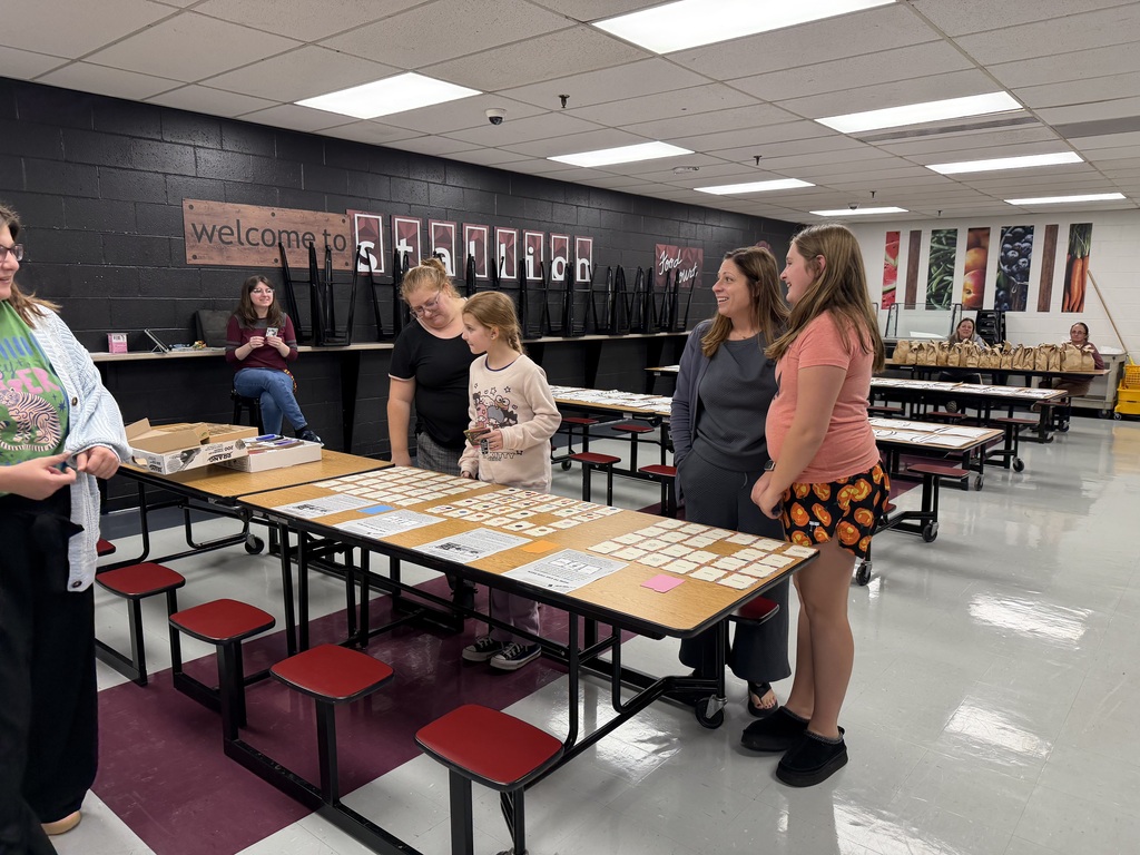 A photo of tables in a cafeteria setting. Papers are on the tables and people are standing around the first table. Some appear to be talking others are looking at things on the table. In the background two adults sit at the last table that has brown bags on it.l