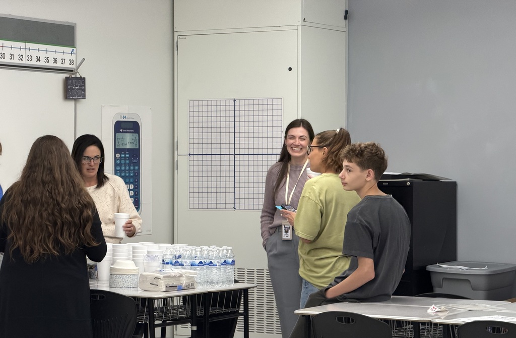 A photo of 5 people standing in a classroom. A teenagers is leaning against a desk and the others appear to be talking. There is a desk covered with a stack of bowls, napkins, and cups. One adult has a cup in her hand.