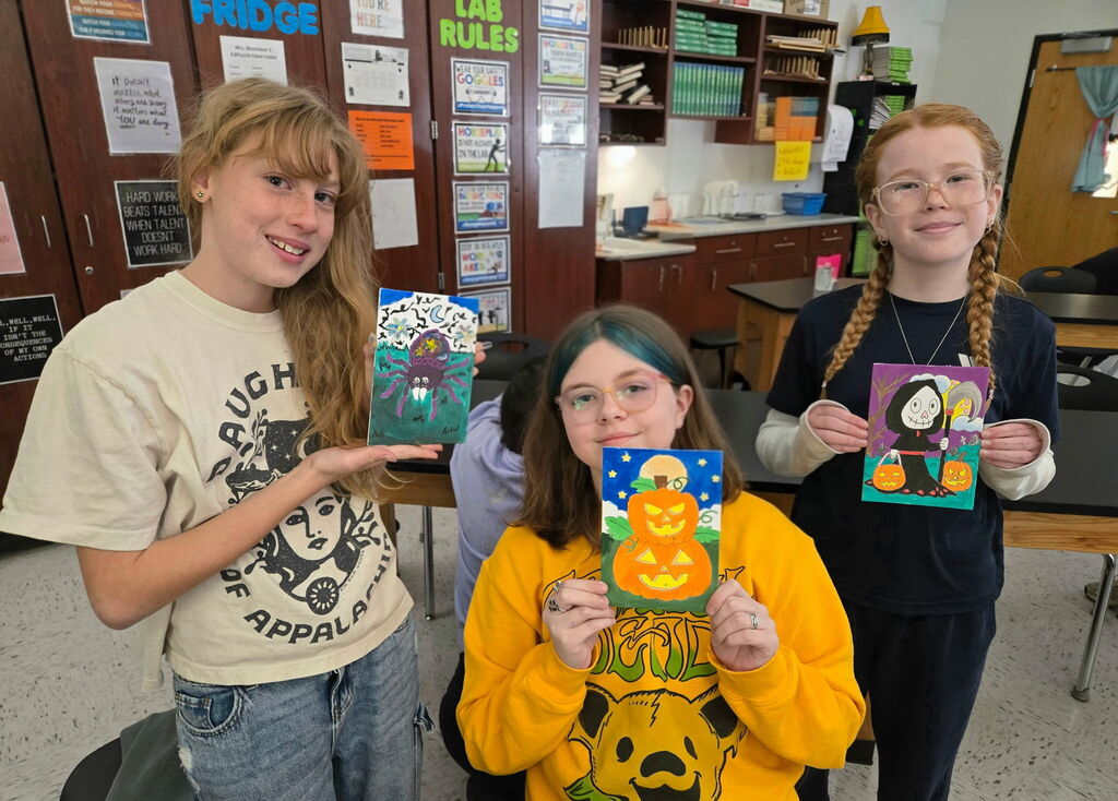 A photo of 3 girls holding Halloween painted pictures. They are in a science classroom.
