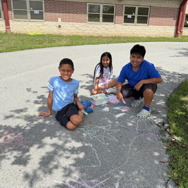 three students sitting on sidewalk with chalk 