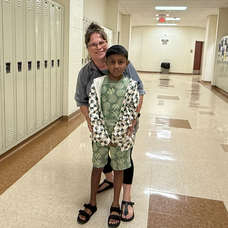 student wearing green shirt and shorts standing with teacher wearing grey shirt in a brown hallway