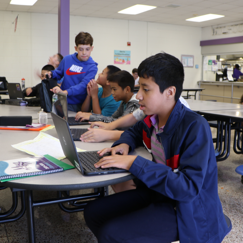 students wearing blue and grey sweatshirts sitting in a lunchroom with black computers