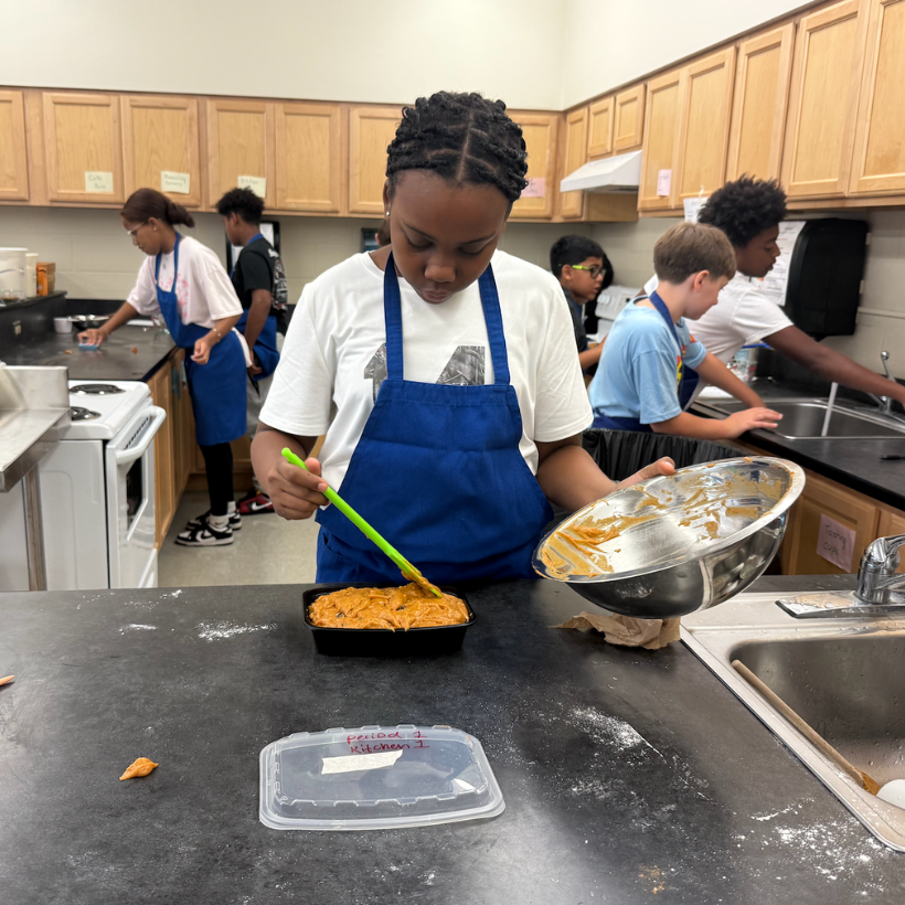 student wearing blue apron pouring mixture 