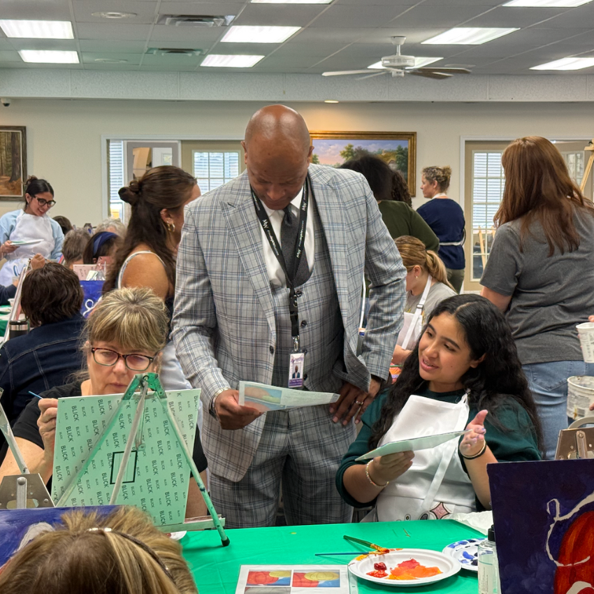 man with a suit standing next to a green table with students holding a paint brush 
