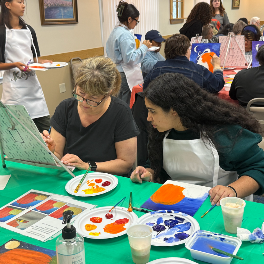 two women with green shirts sitting at a green table with paint brushes 