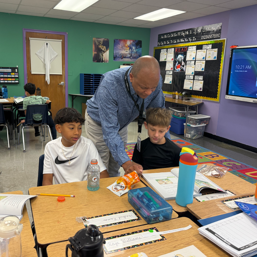 man with a blue shirt helping two students read in a classroom with blue and green walls 