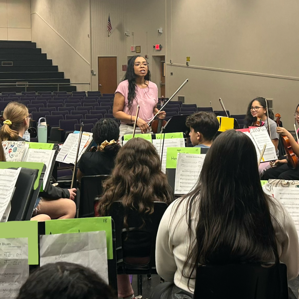 students sitting with violin strings, woman standing talking 