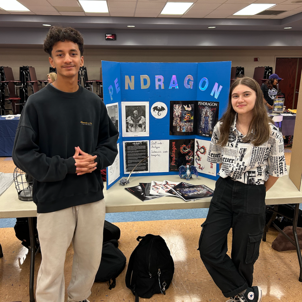 two students smiling standing in front of a poster board that says pendragon