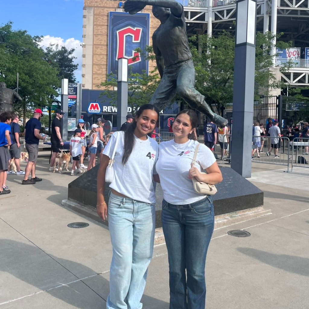 Two women stand before a baseball player statue at a stadium, wearing white shirts and jeans.