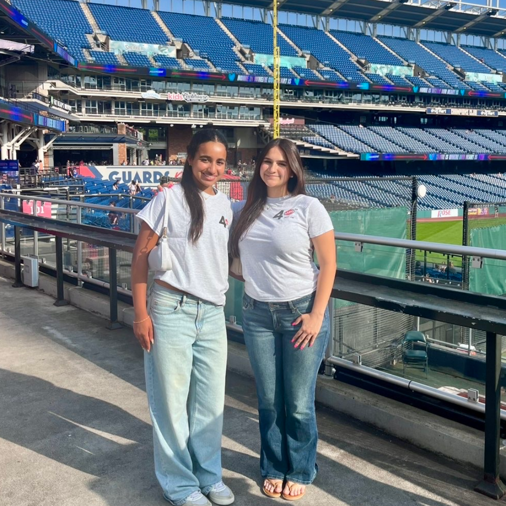 Two women in light-colored shirts stand side by side in a stadium, with empty seats and a railing.