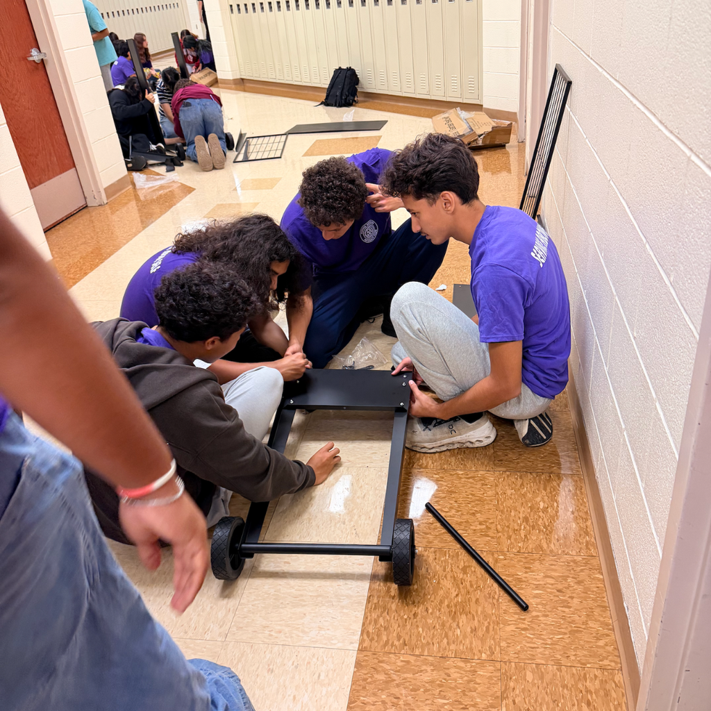Four individuals in matching purple shirts work on a black platform with wheels, in a tiled corridor.