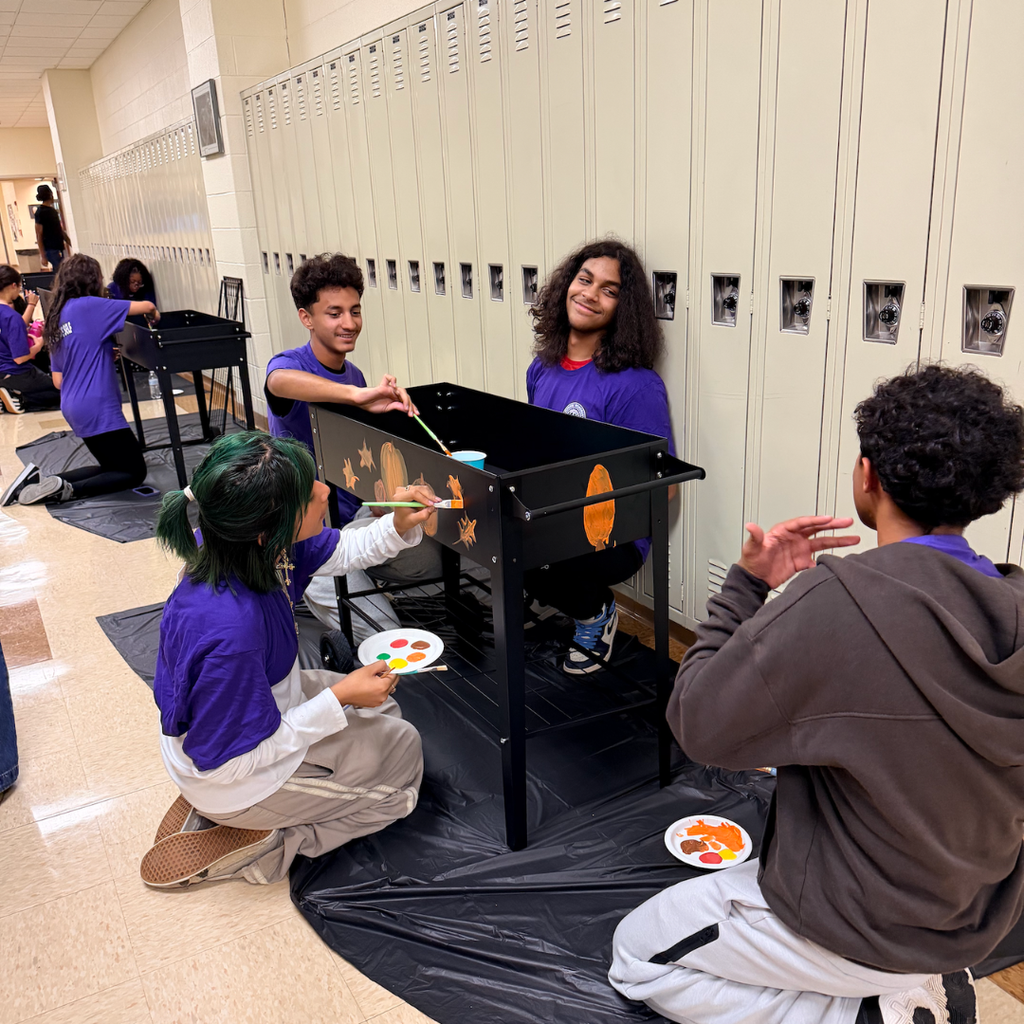 Five individuals, all in purple shirts, painting a black table in a hallway with lockers in the background.