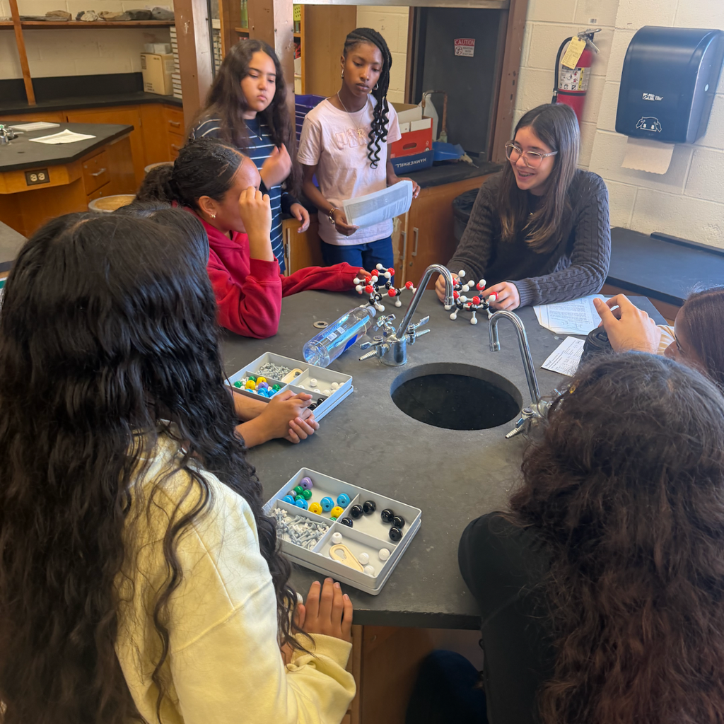 Students in a lab setting, seated around a table with molecular models, a sink, and other materials.