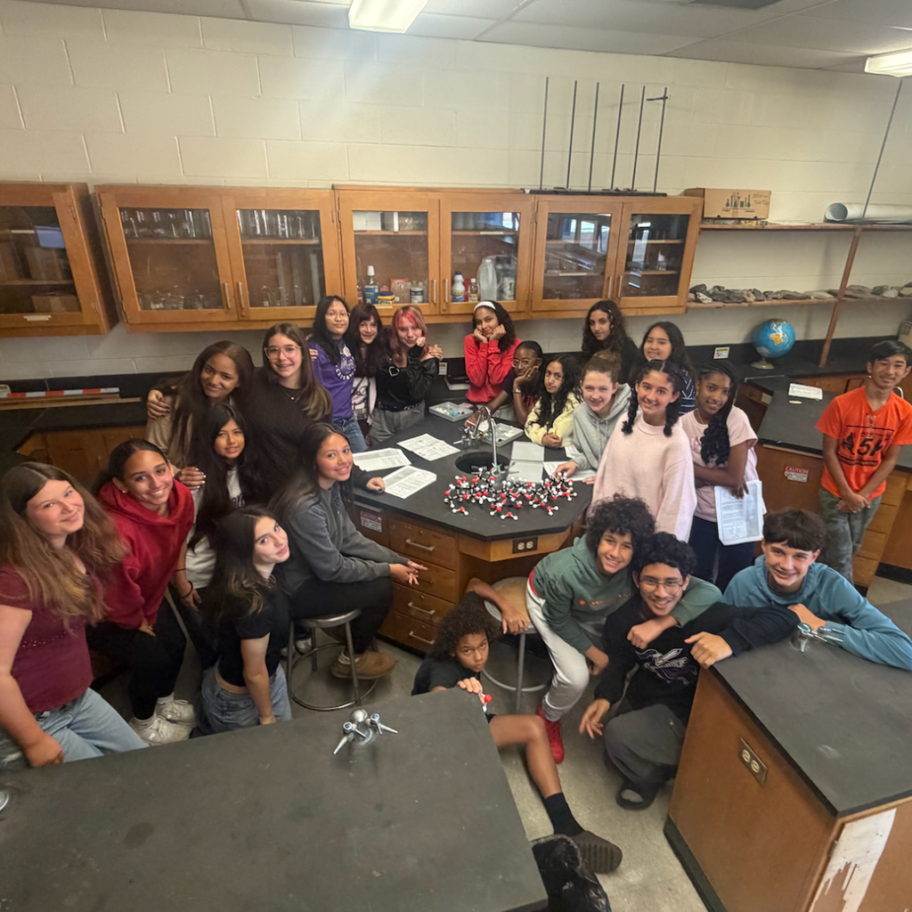A classroom with students, some seated and some standing, posing around a lab station.